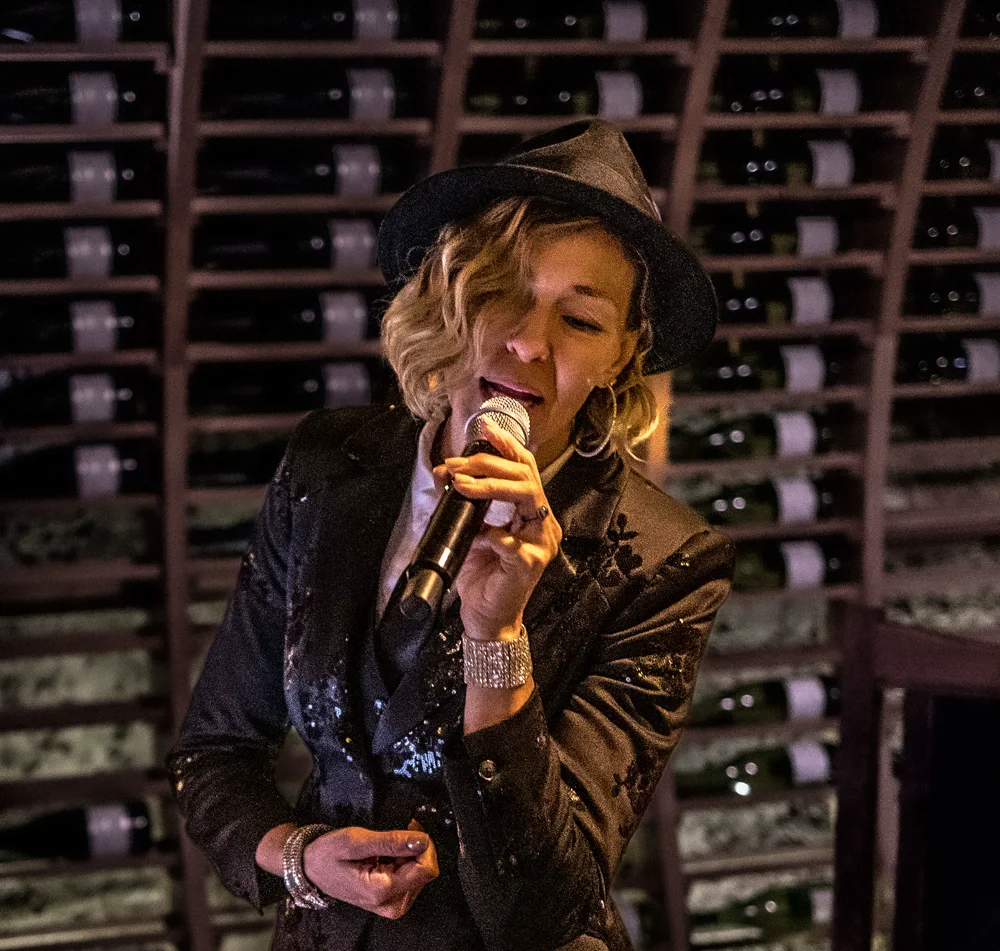 Denise Choate singing in a black hat in front of wine bottle racks